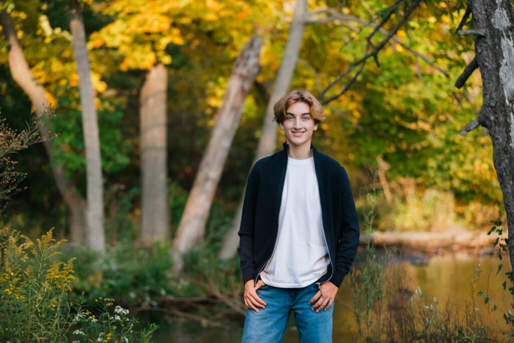 teen boy standing outside near river and trees in autumn